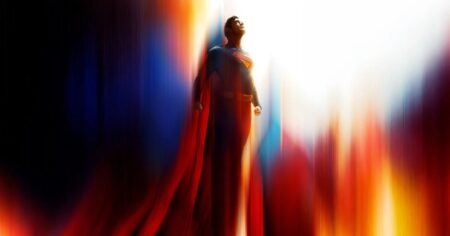 Superman em pé, usando traje azul com capa vermelha, observa o céu iluminado por feixes de luz coloridos em imagem promocional do filme dirigido por James Gunn.
