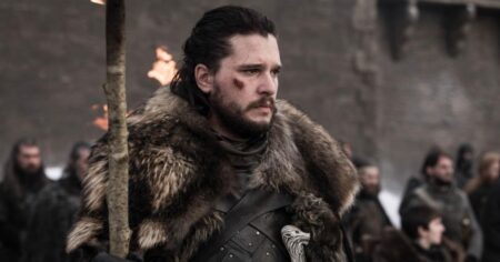 Kit Harington caracterizado como Jon Snow em cena de batalha da série Game of Thrones, usando armadura escura e manto de pele em ambiente medieval coberto de neve