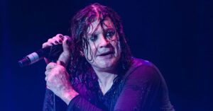 Ozzy Osbourne se apresenta ao vivo no palco, segurando o microfone durante show, com cabelos molhados e iluminação roxa ao fundo