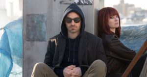 Demolidor: Renascido ganha trailer da 2ª temporada e revela retorno de Jessica Jones Personagens de Demolidor: Renascido sentados em cenário urbano, com homem encapuzado e de óculos escuros em primeiro plano e mulher de cabelo ruivo ao fundo, em clima sombrio e tenso.