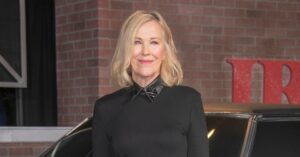Catherine O’Hara posa para fotógrafos em evento de cinema, usando vestido preto, diante de carro clássico