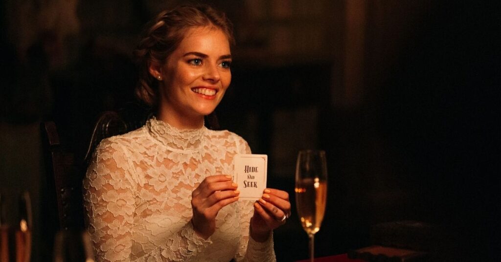 Slashers modernos: 10 filmes de 2010 a 2025 que reinventaram o terror Mulher sorridente vestida de branco segura uma carta com a frase ‘hide and seek’ em um ambiente escuro e elegante, com taça de champanhe ao fundo.
