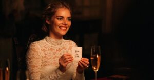 Slashers modernos: 10 filmes de 2010 a 2025 que reinventaram o terror Mulher sorridente vestida de branco segura uma carta com a frase ‘hide and seek’ em um ambiente escuro e elegante, com taça de champanhe ao fundo.