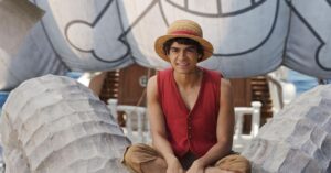 Ator Iñaki Godoy caracterizado como Monkey D. Luffy, usando seu icônico chapéu de palha e colete vermelho, sentado sorrindo na cabeça do navio Going Merry em cena da segunda temporada de One Piece da Netflix.