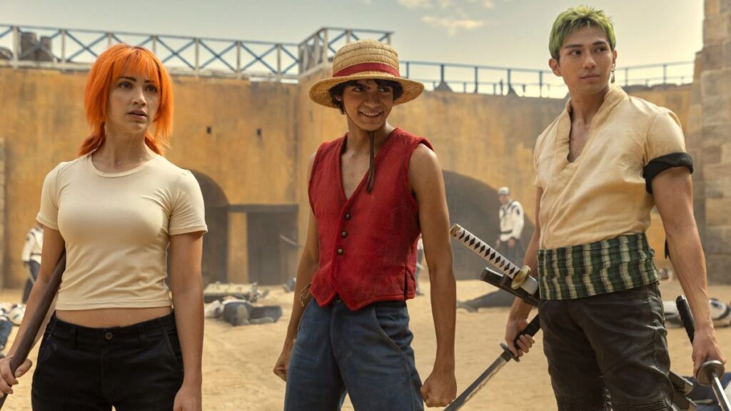One Piece segunda temporada: tudo o que sabemos até agora sobre a nova fase da série Monkey D. Luffy, Nami e Roronoa Zoro em cena do live-action de One Piece, em um pátio de areia após uma batalha; Luffy usa chapéu de palha e colete vermelho, Nami segura um bastão e Zoro empunha uma katana, com guardas caídos ao fundo.