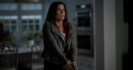 Pânico 7: trailer do Super Bowl sela destino de Sidney Prescott Neve Campbell, como Sidney Prescott, segura uma arma com expressão de alerta em uma cozinha clara durante cena de Pânico 7.