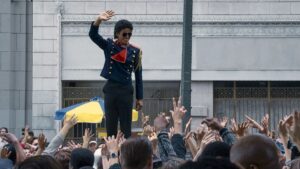 Cinebiografia de Michael Jackson tem duração revelada; confira Jaafar Jackson aparece caracterizado como Michael Jackson, usando uniforme escuro com detalhes vermelhos e óculos escuros, acenando para uma multidão com as mãos erguidas em frente a um prédio.