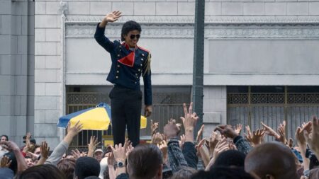 Jaafar Jackson aparece caracterizado como Michael Jackson, usando uniforme escuro com detalhes vermelhos e óculos escuros, acenando para uma multidão com as mãos erguidas em frente a um prédio.