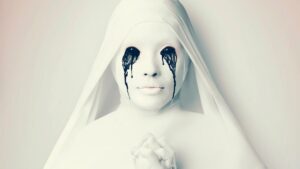 Figura com aparência fantasmagórica vestida com véu branco, rosto totalmente pálido e olhos escuros dos quais escorre uma substância preta semelhante a lágrimas, com as mãos juntas em posição de oração, criando atmosfera sombria e de terror.