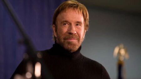 Chuck Norris aparece em close usando suéter preto de gola alta durante um evento, com expressão serena e iluminação suave ao fundo.