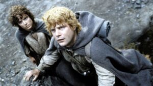Frodo Bolseiro e Samwise Gamgee aparecem escalando uma encosta rochosa em cena do filme O Senhor dos Anéis: O Retorno do Rei, usando capas de viagem enquanto observam algo acima.