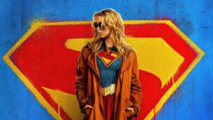 Supergirl: James Gunn revela onde o longa se encaixa na cronologia do novo DCU Supergirl aparece usando uniforme com o símbolo do “S” no peito e sobretudo marrom, posando diante de um grande emblema do Superman grafitado em fundo azul.