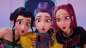 Cena da animação Guerreiras do K‑Pop mostra três cantoras de um grupo idol olhando surpresas para a tela de um celular, reunidas lado a lado em um ambiente colorido inspirado no universo do K-pop.