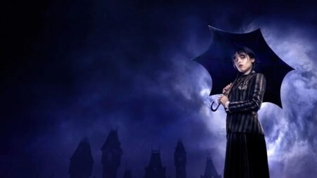 Wednesday Addams aparece vestida com uniforme escuro e segurando um guarda-chuva preto enquanto observa o horizonte sob chuva, tendo ao fundo a silhueta gótica da Academia Nevermore na série Wednesday.