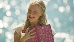 Amanda Seyfried como Sophie em Mamma Mia, sorridente ao ar livre segura um diário rosa com estampa floral, tendo o mar desfocado ao fundo em dia ensolarado.