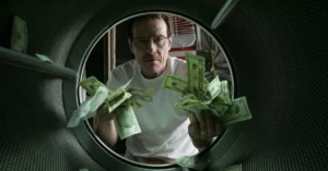 Walter White segurando notas de dinheiro dentro de uma máquina de lavar em cena da série Breaking Bad