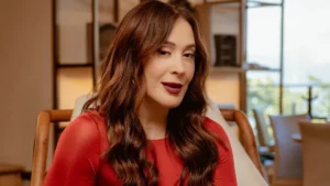Claudia Raia com cabelo ondulado castanho e vestido vermelho posa sentada em ambiente interno iluminado com estantes ao fundo