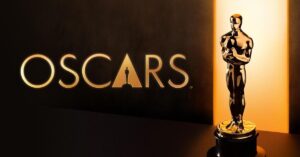 Estatueta dourada do Oscar posicionada ao lado do logotipo “Oscars”, com iluminação em tons quentes destacando o troféu oficial da Academia de Artes e Ciências Cinematográficas - Como funciona o Oscar