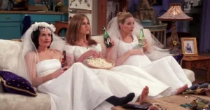 Três personagens de Friends aparecem sentadas em um sofá, vestidas de noiva, enquanto assistem à televisão em cena da sitcom