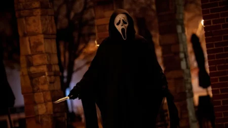 Pânico 7: final explicado, elenco, críticas e onde assistir ao filme Pessoa usando máscara de Ghostface segurando uma faca em um ambiente escuro, com iluminação urbana ao fundo, remetendo a cenas de suspense e terror.