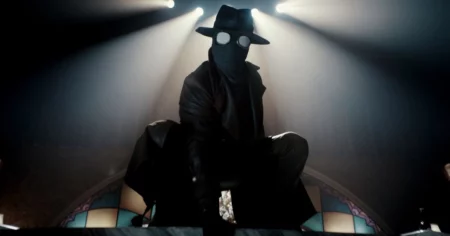 Nicolas Cage caracterizado como Spider-Noir com traje escuro, máscara e chapéu sob luz dramática em ambiente inspirado nos anos 1930