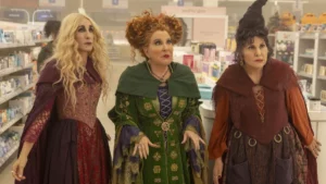 As irmãs Sanderson aparecem em uma farmácia no filme “Abracadabra 2”, usando seus trajes clássicos de bruxas enquanto observam algo com expressão de surpresa.