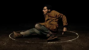 ‘Hokum: O Pesadelo da Bruxa’ ganha novo trailer com astro de ‘Ruptura’; confira Adam Scott aparece assustado dentro de um círculo desenhado no chão em cena sombria de “Hokum: O Pesadelo da Bruxa”.