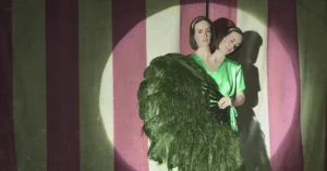 Cena de American Horror Story Freak Show com gêmeas siamesas diante de fundo listrado e iluminação de palco; destaque em texto sobre onde assistir american horror story