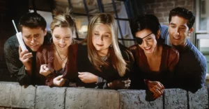 Joey, Phoebe, Rachel, Monica e Ross aparecem juntos apoiados em uma sacada durante cena da série Friends.