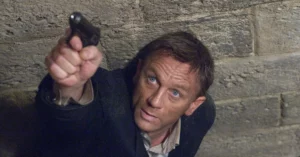 Daniel Craig como James Bond aponta arma em cena de ação da franquia 007, diante de uma parede de pedra