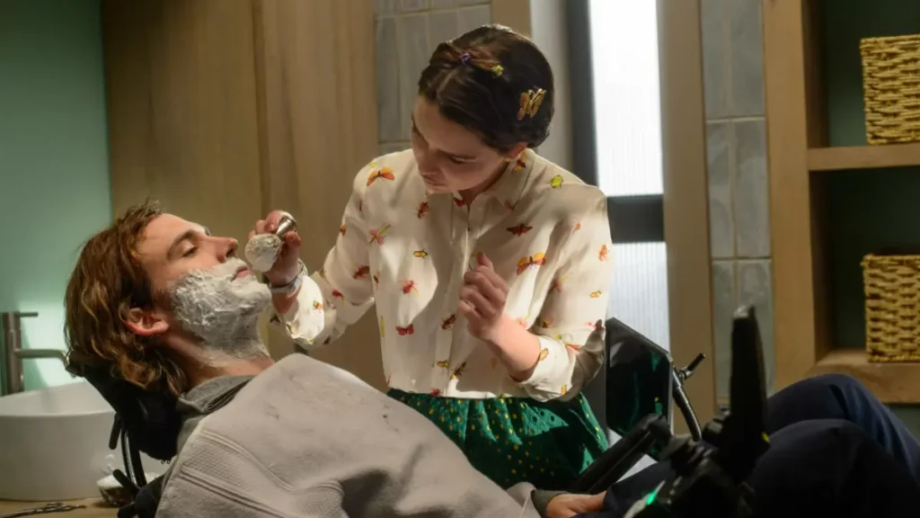 Uma cena do filme Como Eu Era Antes de Você, mostrando os personagens Louisa Clark (Emilia Clarke) e Will Traynor (Sam Claflin) em um banheiro iluminado. Will está reclinado em sua cadeira de rodas motorizada, usando uma toalha cinza protetora sobre o peito, com o rosto coberto por espuma de barbear branca. Louisa está de pé ao lado dele, inclinando-se ligeiramente para a frente concentrada. Ela usa uma camisa branca de botões com estampas coloridas de borboletas e insetos, uma saia verde e presilhas de borboleta no cabelo preso. Ela segura um pincel de barbear de metal na mão direita, aplicando a espuma nas bochechas de Will. Ao fundo, uma pia branca e prateleiras de madeira são visíveis.