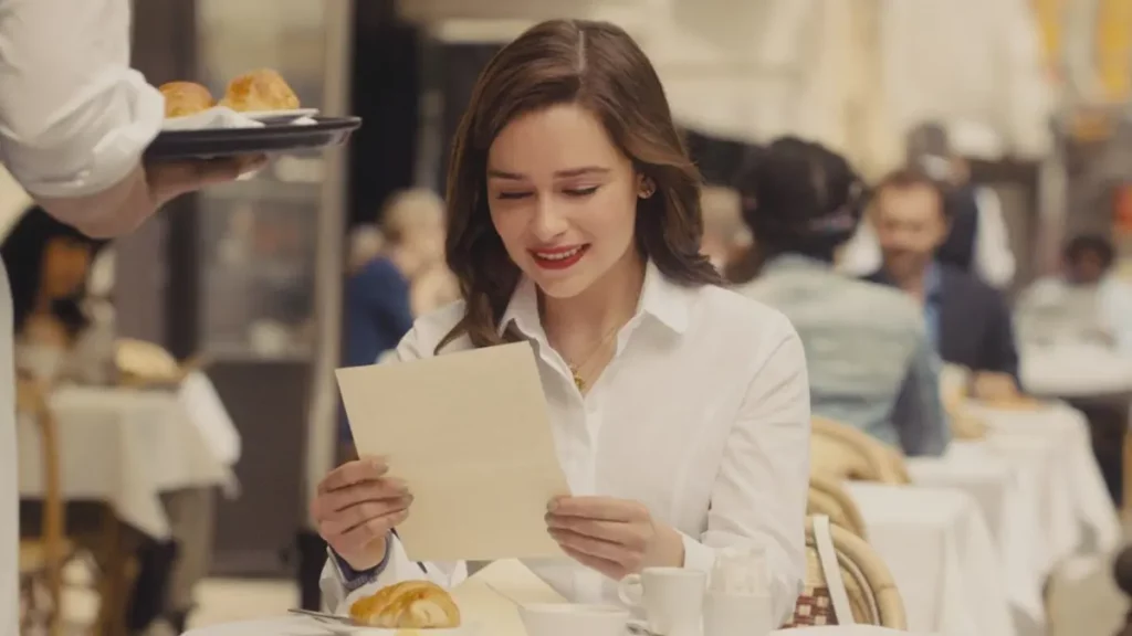 Cena do filme Como Eu Era Antes de Você, destacando a personagem Louisa Clark (Emilia Clarke) sentada em um café ao ar livre em Paris. Ela está vestindo uma camisa social branca de botões, usa batom vermelho marcante e tem um olhar baixo e um sorriso suave e emocionado enquanto lê uma carta que segura com as duas mãos. 