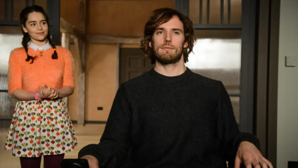 Cena do filme Como Eu Era Antes de Você, com o personagem Will Traynor (Sam Claflin) em primeiro plano e Louisa Clark (Emilia Clarke) ao fundo. Will está sentado em sua cadeira de rodas, olhando diretamente para a frente com uma expressão séria e pensativa. Ele tem cabelos castanhos ondulados, barba e veste um suéter de tricô cinza-escuro. Atrás dele, ligeiramente à esquerda, Louisa está de pé com as mãos timidamente unidas e um sorriso discreto. Ela usa um suéter felpudo laranja-vivo sobre uma camisa de gola branca, uma saia curta estampada com pequenos rabanetes e meia-calça escura. O cabelo dela está preso em duas tranças laterais com elásticos coloridos.
