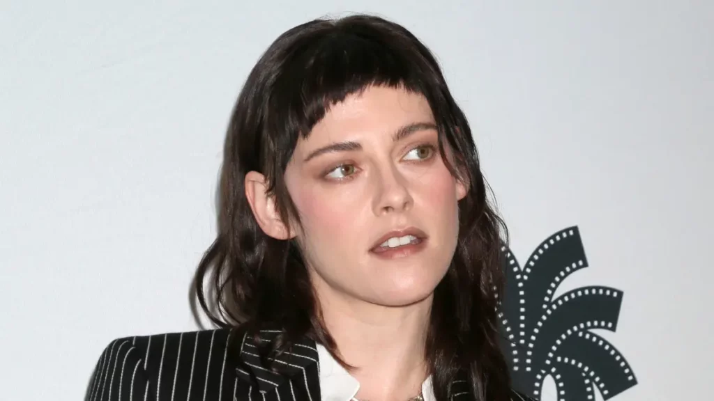 Kristen Stewart abre o jogo sobre filme com Wagner Moura: ‘pesadelo magnífico’ Foto de rosto e ombros de Kristen Stewart, com expressão séria e olhando para a direita. Ela usa um terno risca de giz preto e branco sobre uma camisa branca. O cabelo castanho-escuro está na altura dos ombros, e ela tem uma franja curtíssima (estilo "baby bangs"). O fundo é branco, com uma logo de um festival de cinema (formato de folha de palmeira com perfurações de filme) em relevo no lado direito.