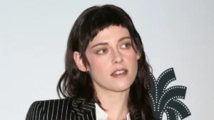 Foto de rosto e ombros de Kristen Stewart, com expressão séria e olhando para a direita. Ela usa um terno risca de giz preto e branco sobre uma camisa branca. O cabelo castanho-escuro está na altura dos ombros, e ela tem uma franja curtíssima (estilo "baby bangs"). O fundo é branco, com uma logo de um festival de cinema (formato de folha de palmeira com perfurações de filme) em relevo no lado direito.