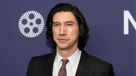 Adam Driver posa para fotógrafos usando terno escuro, camisa branca e gravata durante um evento.