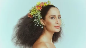 Camila Pitanga será Clara Nunes em cinebiografia sobre a sambista; confira Camila Pitanga caracterizada como Clara Nunes. Mulher em perfil com cabelo volumoso adornado por flores coloridas, olhando para a câmera, sobre fundo azul-claro.