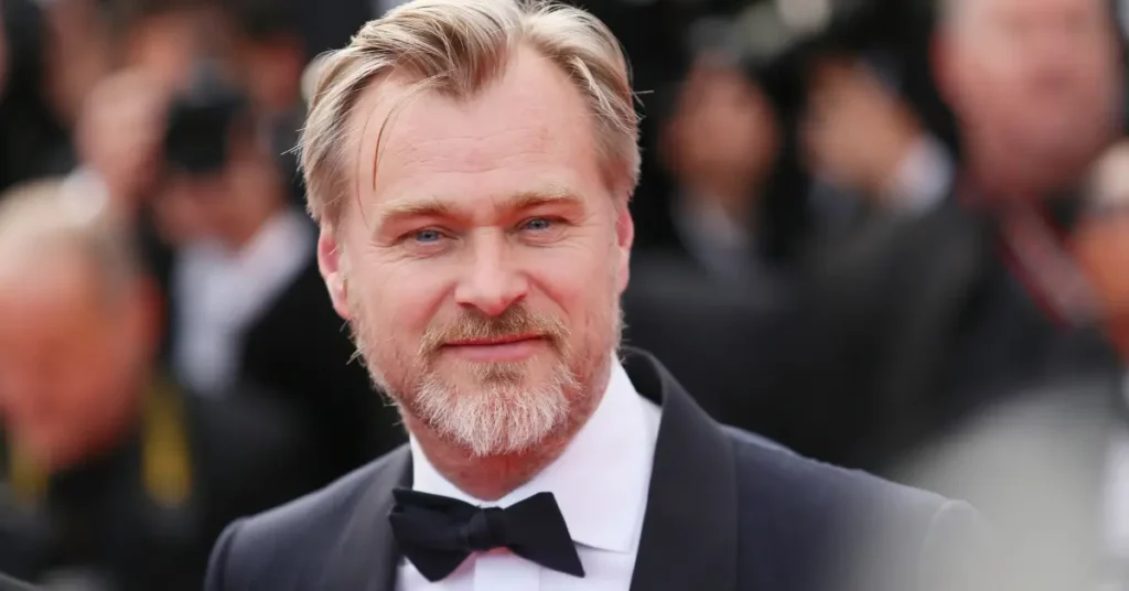 Christopher Nolan revela receio de dirigir comédia romântica: ‘seria difícil’ Christopher Nolan, homem de barba grisalha e terno preto com gravata borboleta, sorri para a câmera em evento de tapete vermelho.