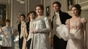 Elenco de Bridgerton: conheça os atores e personagens da série Sete membros do elenco de Bridgerton alinhados em um luxuoso salão. A matriarca Violet Bridgerton está em destaque ao centro com um vestido prateado, cercada por alguns de seus filhos e sua nora Kate Sharma, todos vestindo trajes de época formais e observando algo com expressões atentas.