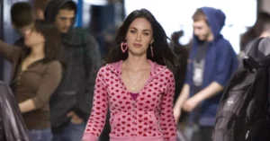 Megan Fox como Jennifer Check caminha por um corredor usando casaco rosa estampado com corações em cena de Garota Infernal