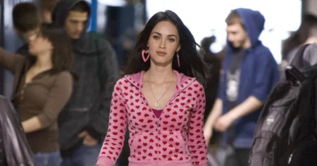 Megan Fox como Jennifer Check caminha por um corredor usando casaco rosa estampado com corações em cena de Garota Infernal