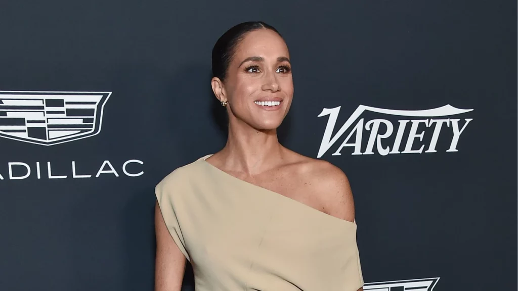 Meghan Markle sorri durante evento de gala, usando vestido bege de um ombro só, diante de um painel com logotipos da Variety e da Cadillac.