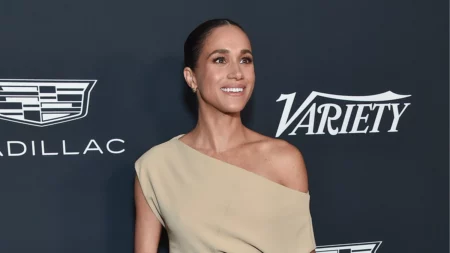Meghan Markle sorri durante evento de gala, usando vestido bege de um ombro só, diante de um painel com logotipos da Variety e da Cadillac.