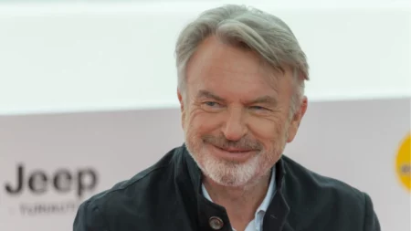 Sam Neill será visto em 3 filmes após a morte; veja quais são Sam Neill sorri durante uma aparição pública usando casaco preto sobre camisa clara.