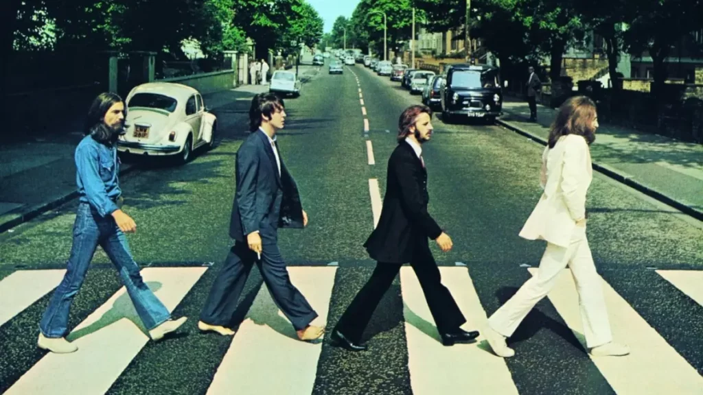 Beatles: atores recriam capa de ‘Abbey Road’ e flagra de bastidores impressiona fãs Fotografia colorida da capa do álbum Abbey Road. Os quatro integrantes da banda Beatles atravessam uma faixa de pedestres em fila indiana, em uma rua arborizada com carros estacionados ao fundo.