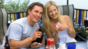 Fotografia de Matthew McConaughey e Kate Hudson sorrindo e se divertindo juntos enquanto estão sentados a uma mesa ao ar livre. Kate segura um baralho vermelho e Matthew aponta para a frente.