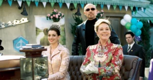 Anne Hathaway abre o jogo sobre Julie Andrews em O Diário da Princesa 3 Anne Hathaway aparece à esquerda usando tiara e roupa rosa, enquanto Julie Andrews está sentada à direita com traje floral vermelho e branco em cena de O Diário da Princesa 2. Ao fundo, seguranças acompanham as personagens em um ambiente externo decorado com bandeirolas e balões