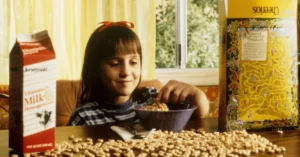 Matilda faz 30 anos; veja o antes e depois do elenco Matilda com cabelos escuros sentada à mesa diante de uma tigela de cereal, com caixas de leite e sucrilhos ao redor