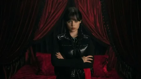 Fotografia de Jenna Ortega caracterizada como a protagonista da série, com expressão séria, cabelos trançados e braços cruzados, vestindo preto em um quarto com cortinas e cama de veludo vermelho escuro. A imagem ilustra o retorno da personagem na aguardada Wandinha 3ª temporada.