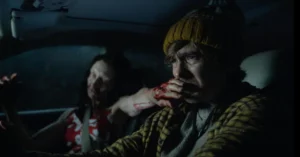 Homem dirige um carro em cena tensa do novo filme Resident Evil enquanto uma criatura aparece ao seu lado
